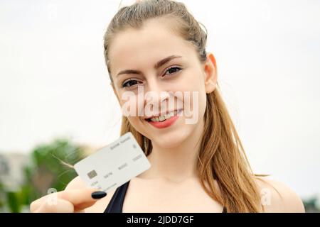 Jeune femme souriante confiante portant un soutien-gorge de sport debout sur le parc de la ville, à l'extérieur montrant une carte de crédit ou de débit en plastique, effectuant des paiements en ligne. Sélection Banque D'Images