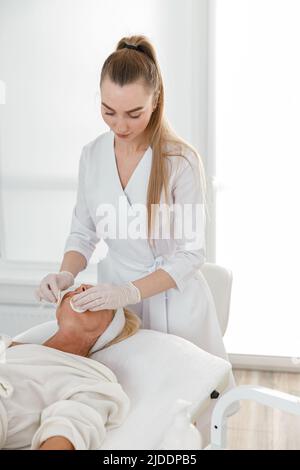 Belle femme médecin professionnel nettoie une peau de femme mûre dans un centre moderne de cosmétologie de lumière Banque D'Images