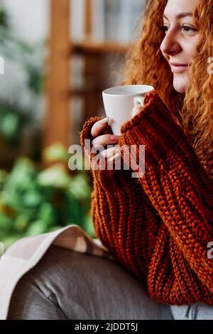 Une petite fille de gingembre mignon aux cheveux bouclés est assise dans la chaise à la maison le matin et boit son café. Banque D'Images