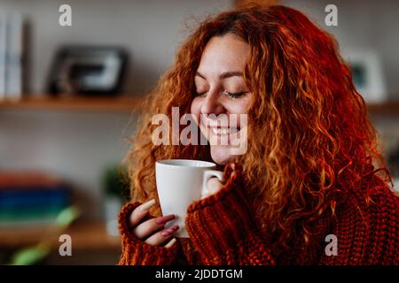 Une petite fille de gingembre mignon aux cheveux bouclés est assise dans la chaise à la maison le matin et boit son café. Banque D'Images