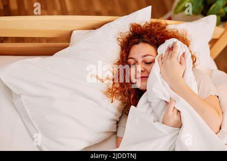 Paresseuse fille à tête rouge dans le lit. Une petite fille de gingembre paresseuse couchée dans son lit le matin, couvrant sa tête avec un drap de lit, et ne veut pas se lever. Banque D'Images