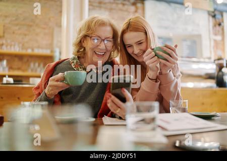 Une grand-mère heureuse et sa petite-fille adolescente assise dans un café, en dégustant un café et en utilisant un smartphone pour des vidéos amusantes. Banque D'Images