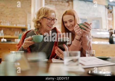 Une grand-mère heureuse et sa petite-fille adolescente assise dans un café, en dégustant un café et en utilisant un smartphone pour des vidéos amusantes. Banque D'Images