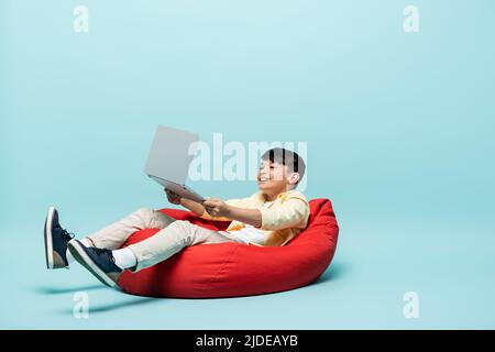 Enfant asiatique positif tenant un ordinateur portable tout en étant assis sur une chaise de sac à bandoulière sur fond bleu Banque D'Images