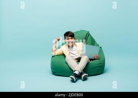 Un écolier asiatique souriant montrant oui geste et tenant un ordinateur portable sur une chaise de sac à bandoulière sur fond bleu Banque D'Images