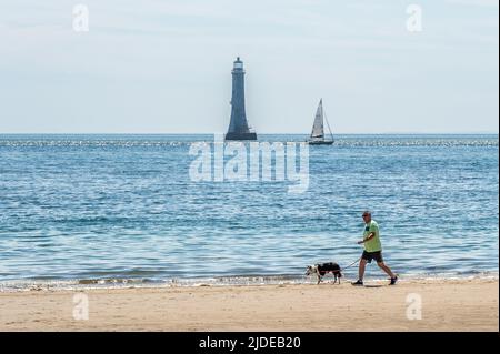 Cranfield, County Down, Royaume-Uni. Lors d'une journée de soleil et de sommets de 20C, de nombreuses personnes ont frappé les plages d'Irlande du Nord. Un homme marche son chien le long du rivage de Cranfield Beach avec le phare de Haulbowline comme toile de fond. Crédit : AG News/Alay Live News Banque D'Images