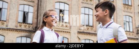 Écoliers multiethniques avec sacs à dos parlant près de l'école floue à l'extérieur, bannière Banque D'Images