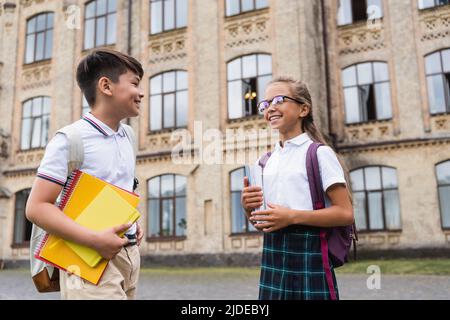 Des élèves gais et interraciaux avec des carnets qui parlent près d'une école floue à l'extérieur Banque D'Images