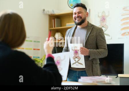 Joyeux enseignant de sexe masculin avec une affiche de structure interne de grenouille regardant l'élève la décrivant tout en étant assis devant lui Banque D'Images