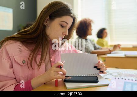 Adolescente en veste de denim rose textant dans un smartphone à la leçon derrière la page de copybook tout en étant assis près du bureau contre ses camarades de classe Banque D'Images