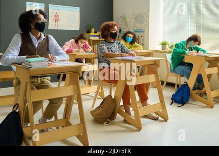 Adolescents interculturels élèves de l'école secondaire dans des masques protecteurs à l'écoute de l'enseignant tout en étant assis près des bureaux à la leçon d'anatomie Banque D'Images