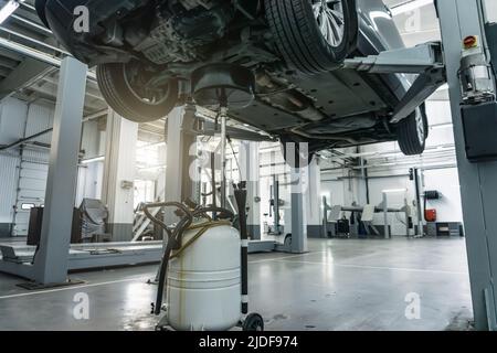 Vidange de l'huile moteur lors de l'entretien de la voiture. Auto est sur le relevage hydraulique dans l'atelier de garage, processus de vidange de l'ancienne huile usagée sombre. Banque D'Images