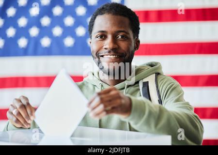 Portrait d'un homme noir souriant mettant le bulletin de vote dans le bac contre le drapeau américain Banque D'Images