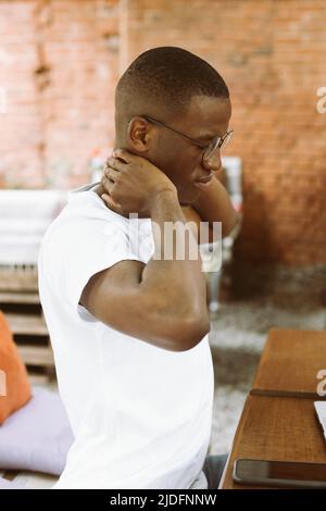 Vertical afro-américain, homme de peau sombre en lunettes, assis avec un ordinateur portable dans un café de rue, massant son cou. Travail sédentaire Banque D'Images