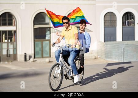 Jeune couple gay tenant un drapeau de fierté gay tout en faisant un vélo à l'extérieur. LGBT, relation et égalité des droits. Banque D'Images