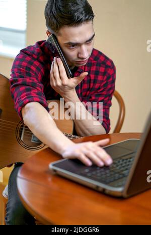 Le jeune homme apprend à jouer de la guitare, prend des leçons en utilisant l'Internet se trouve près d'une table basse avec une tasse de thé chaud . Poste de travail près de la fenêtre. Travail Banque D'Images