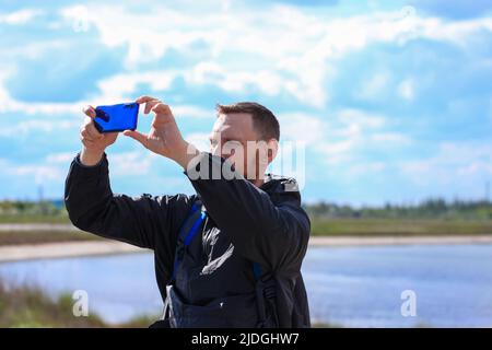 Homme barbu en imperméable noir sur fond bleu de paysage de mer avec smartphone entre les mains prend la photographie de selfie. Portrait de la course à pied de jeunes vêtements taille basse Banque D'Images
