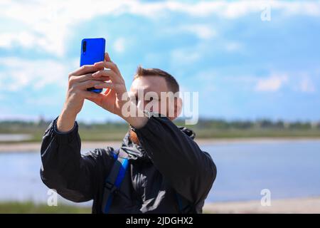 Homme barbu en imperméable noir sur fond bleu de paysage de mer avec smartphone entre les mains prend la photographie de selfie. Portrait de la course à pied de jeunes vêtements taille basse Banque D'Images