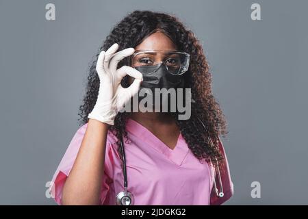 Jeune femme afro-américaine médecin en gants masque et lunettes sur fond gris souriant détendu et gai. Succès et optimisme Banque D'Images