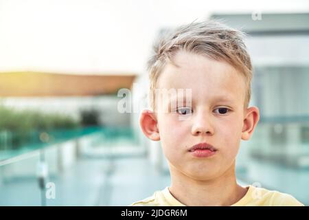 Portrait d'un élève triste avec des cheveux blonds sur un arrière-plan flou. Un petit garçon mignon a l'air de se tenir debout contre le territoire de l'hôtel à proximité de la station Banque D'Images