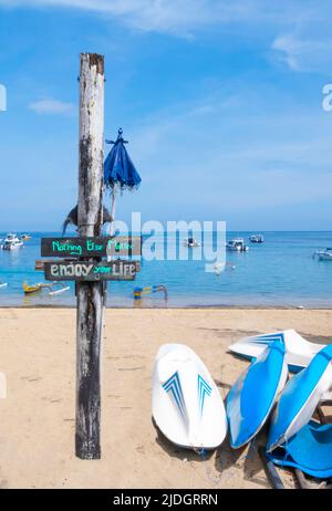 Profitez de votre vie panneau en bois sur le poteau surmonté d'intempéries par des flotteurs d'eau et des planches de surf sur la plage de sable avec des bateaux flottant dans l'océan en arrière-plan. Copier l'espace Banque D'Images