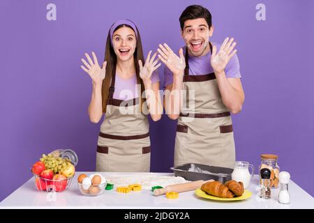 Photo de deux personnes positives excitées lever les bras paumes préparer la pâte isolée sur fond violet Banque D'Images