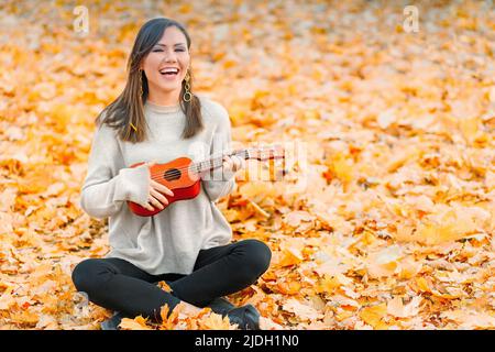 Jeune femme heureuse ayant un bon moment avec un instrument de musique assis sur des feuilles d'automne tombées Banque D'Images