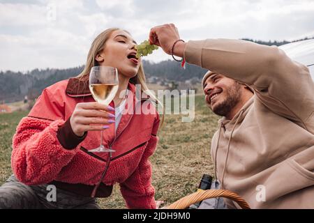 Jeune couple moderne souriant ayant un voyage dans la nature, appréciant le vin et le raisin tout en étant assis détendu à côté de leur nouvelle voiture électrique exclusive Banque D'Images