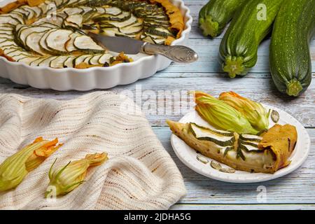 Tarte ouverte aux courges farcies aux herbes et aux caillés de fromage dans un plat de cuisson et sur une assiette sur une table rustique en bois entourée d'ingrédients et de fleurs Banque D'Images