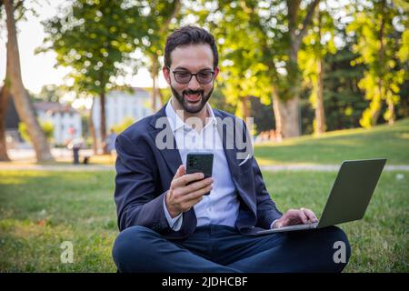 Homme d'affaires assis dans un pré utilise son ordinateur portable et regarde l'écran de son smartphone Banque D'Images