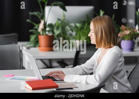 vue latérale d'un directeur publicitaire souriant qui tape sur un ordinateur portable au bureau Banque D'Images