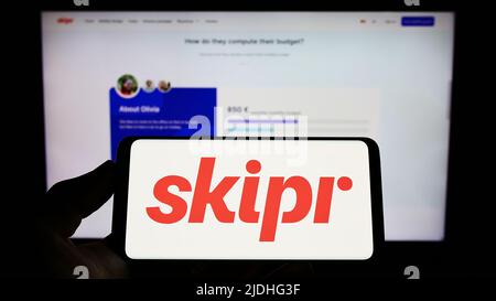 Personne tenant un téléphone portable avec le logo de la société belge de mobilité Skipr sa à l'écran en face de la page web d'affaires. Mise au point sur l'affichage du téléphone. Banque D'Images