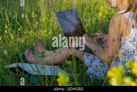 Une petite fille mignonne est assise dans un parc au milieu de fleurs et de types sur un ordinateur portable Banque D'Images