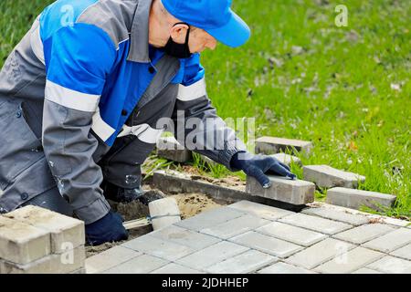 La couche de briques des vêtements de travail se trouve sur le trottoir et pose des dalles de pavage. Vue de l'homme en plein air. Constructeur professionnel fait l'arrangement de territoire jour d'été. Scène réelle. Banque D'Images