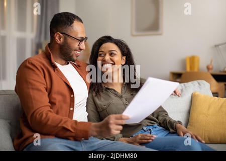 Joyeux couple noir tenant des papiers assis sur un canapé à la maison Banque D'Images