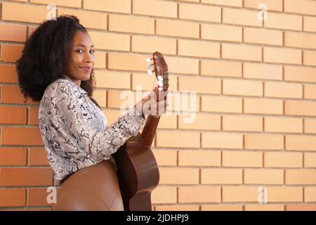 Une jeune femme debout jouant de la guitare penchée contre un mur de briques Banque D'Images
