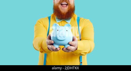 Tirelire en forme de cochon bleu dans les mains d'homme gras barbu joyeux et méconnu. Banque D'Images
