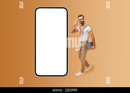 Souriant attrayant gars en lunettes et élégant décontracté porter marcher près d'un immense smartphone avec écran vide, regarder l'appareil photo et sourires, hipster homme annonçant une nouvelle application ou site Web, maquette Banque D'Images