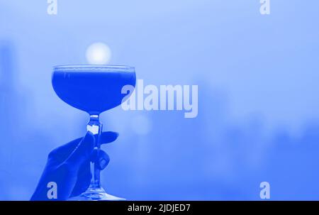 Cocktail en verre bleu cobalt de style pop art à la main avec vue panoramique sur la ville en toile de fond Banque D'Images