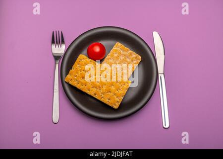 Une seule tomate et une tranche de pain croustillant sur une assiette noire unie et des couverts avec des signes d'utilisation sur fond violet Banque D'Images