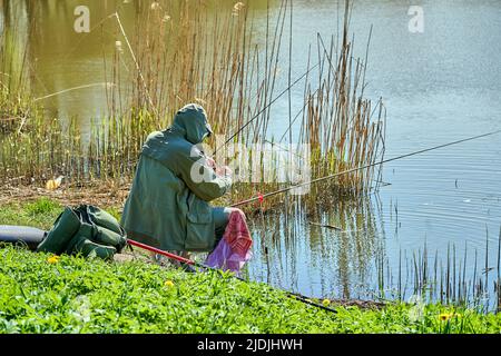 Riga, Lettonie - 8 mai 2022: Un pêcheur est assis avec une canne à pêche sur la rive d'un lac dans un parc public. Herbe verte autour. Vue de l'arrière. Banque D'Images