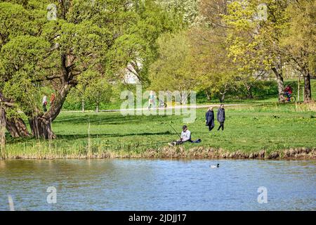 Riga, Lettonie - 8 mai 2022: Un pêcheur est assis avec une canne à pêche sur la rive d'un lac dans un parc public. Herbe verte autour. Les gens marchent à distance. Banque D'Images