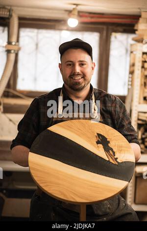 Photo verticale du joiner souriant et heureux en uniforme de protection et capuchon en atelier, tenir la table circulaire. Travail de fabrication Banque D'Images