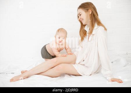 Portrait d'une jeune mère heureuse maman avec de longs cheveux foncés dans une chemise blanche avec un petit mignon petit bébé plumeux assis sur un lit blanc. Banque D'Images