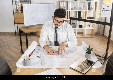 Architecte arabe qualifié en chemise blanche, cravate et lunettes de contrôle projet de construction avec plan et ordinateur portable assis sur une table. Homme musulman séduisant et barbu contrôlant le processus de travail. Banque D'Images