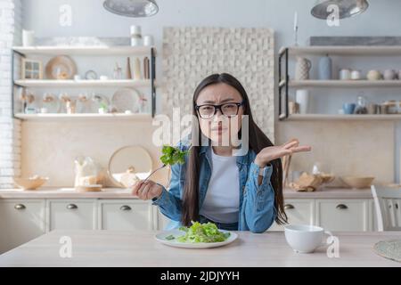 Portrait d'une jeune femme asiatique insatisfaite qui mange de la salade à la maison et regarde l'appareil photo Banque D'Images