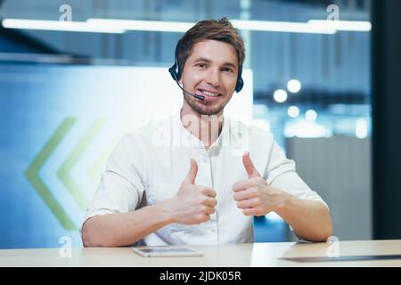 Portrait d'un jeune homme beau dans le casque montre ses doigts super, tient un séminaire en ligne, conférence, regarde la caméra, explique, sourit, dit Banque D'Images
