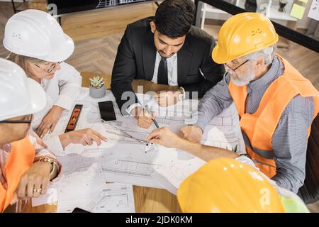 Architectes, ingénieurs et concepteurs qualifiés ayant des réunions d'affaires au bureau. Collègues multiculturels en costumes et casques assis à table et regardant les plans. Concept de construction. Banque D'Images