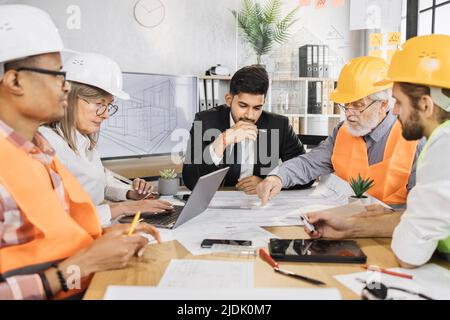 Architectes et ingénieurs en costumes et casques ayant une réunion de travail au bureau moderne. Des collègues de différents âges et de différentes races. Brainstorming d'entreprise. Banque D'Images