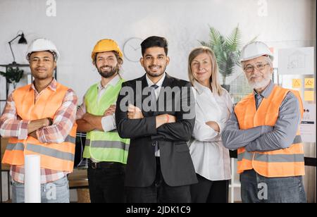 Vue de face d'une femme âgée et de quatre hommes ingénieurs, constructeurs et architectes regardant la caméra tout en se tenant sur un bureau moderne et léger. Personnes d'âge, de sexe et de race différents. Banque D'Images